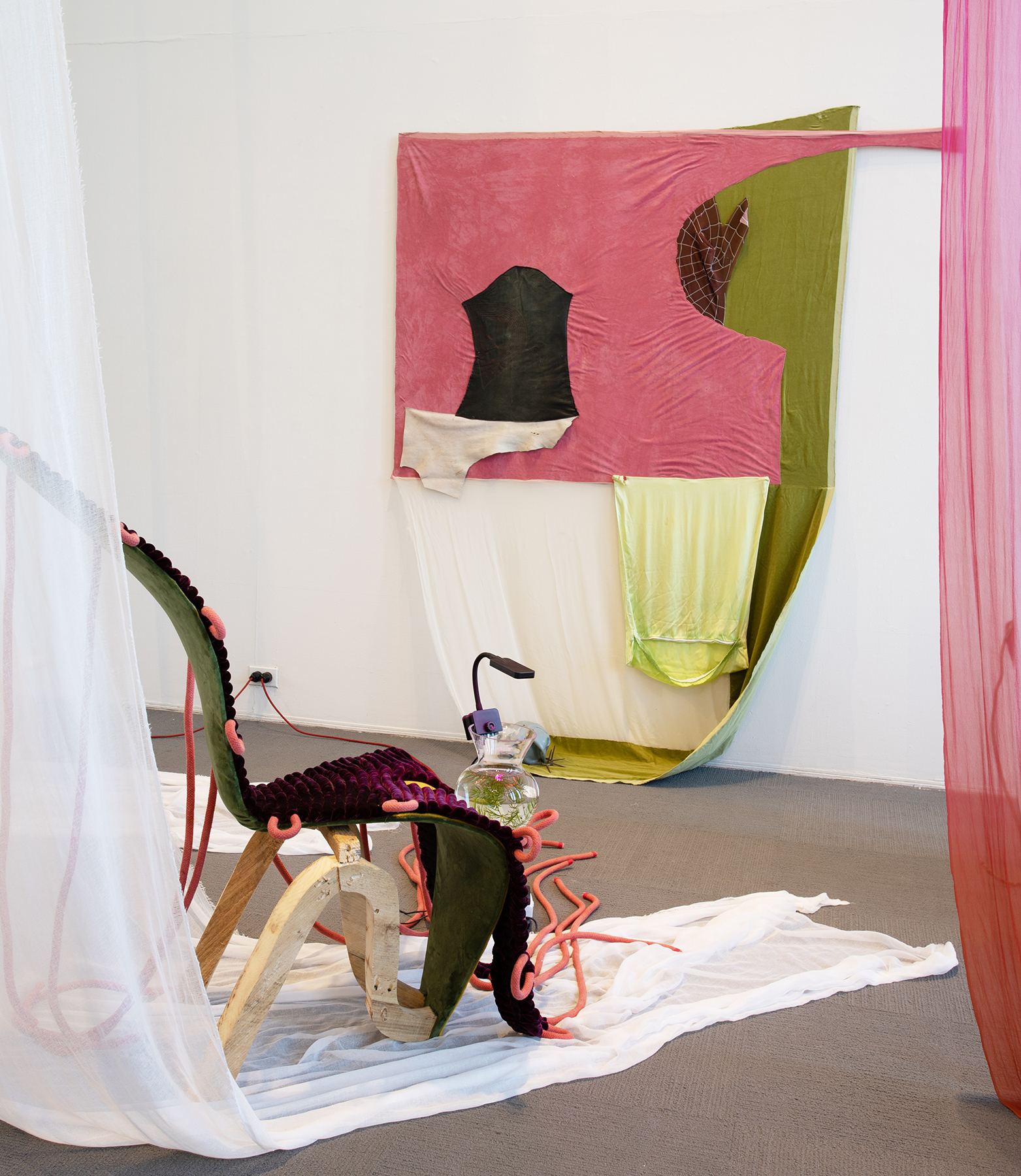 A lounge chair made from lumber and velvet framed by hanging curtains. At the bottom of the seat sits a glass vase with plants. A fabric-covered painting is in the background with elements falling to the floor.