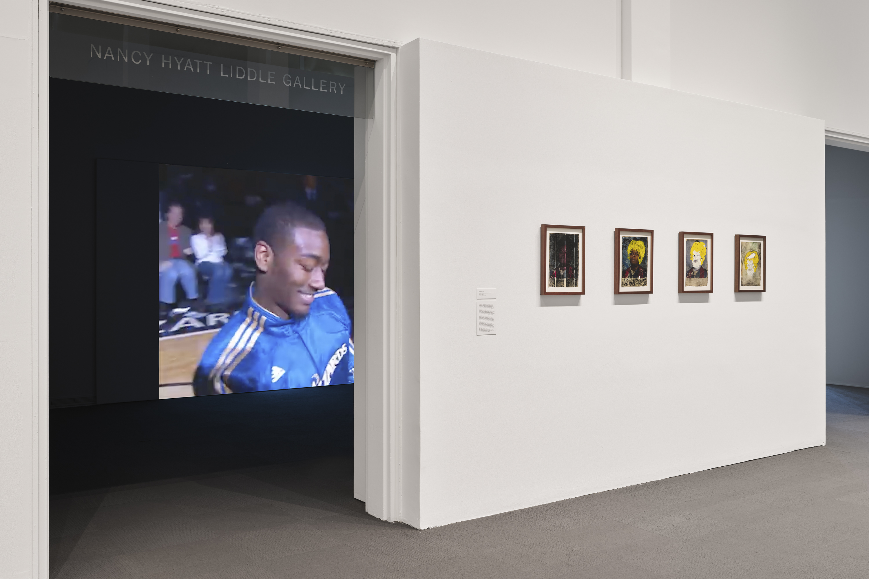 Four framed works on paper installed on a white wall in the University Art Museum. To the left of the wall, there is an opening to the Nancy Hyatt Liddle Gallery, where a video projection by Noel W Anderson and Solomon Bennett is being played. 