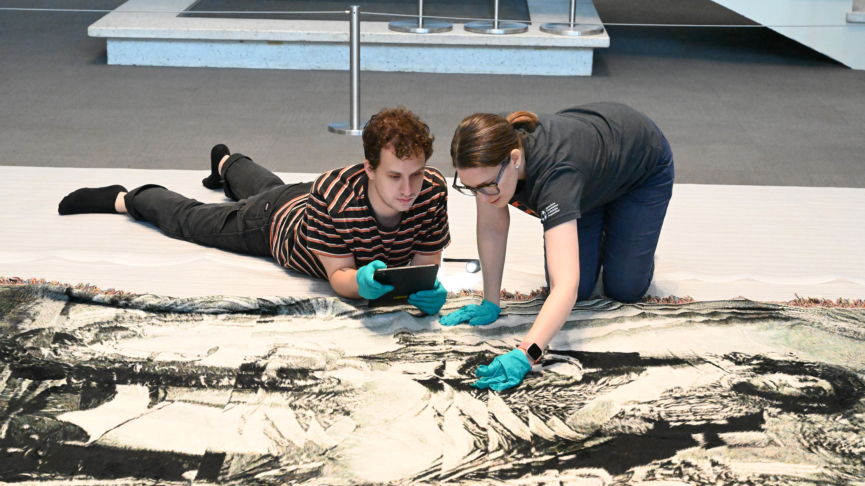 Christian Henry Wechgelaer MFA ’25, Museum Assistant, and Darcie Abbatiello, Registrar/Collections Manager, wear aqua nitrile gloves and closely examine a black and white tapestry by exhibiting artist Noel W Anderson.