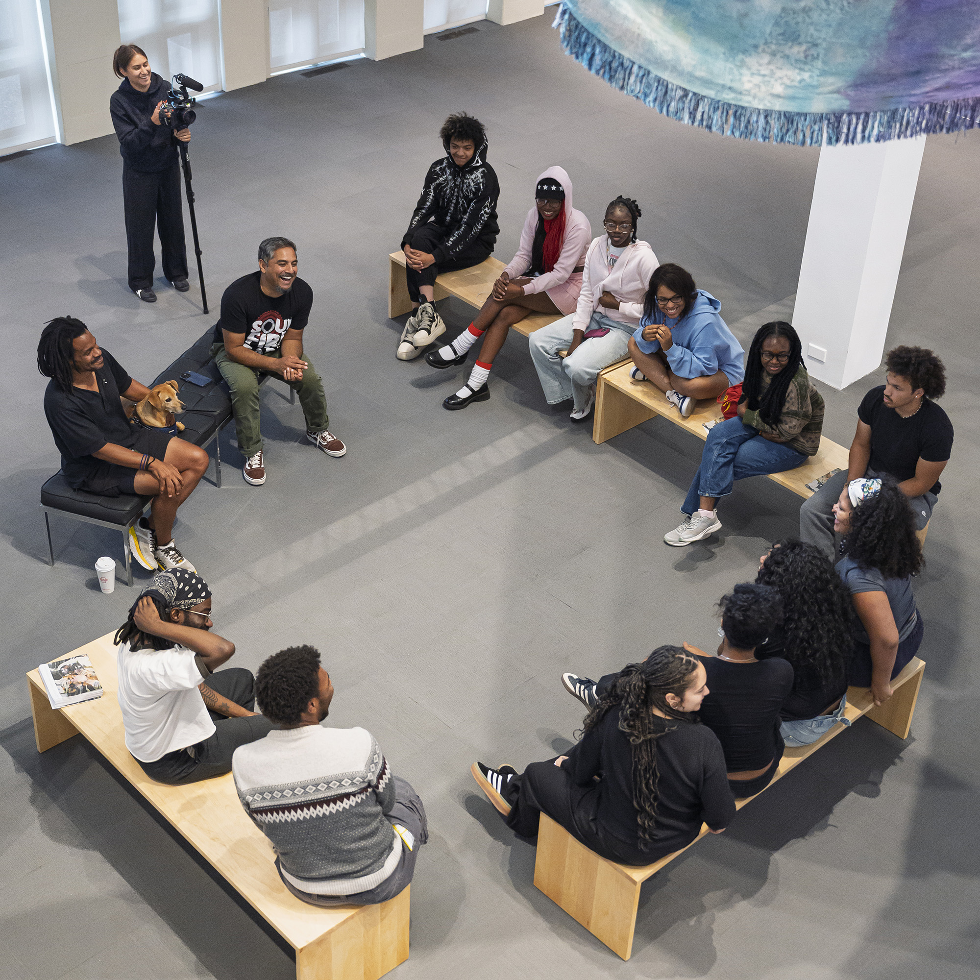 A group of Youth FX students sit on benches arranged in a circle formation in the University Art Museum, engaging in conversation with exhibiting artist Noel W Anderson. 