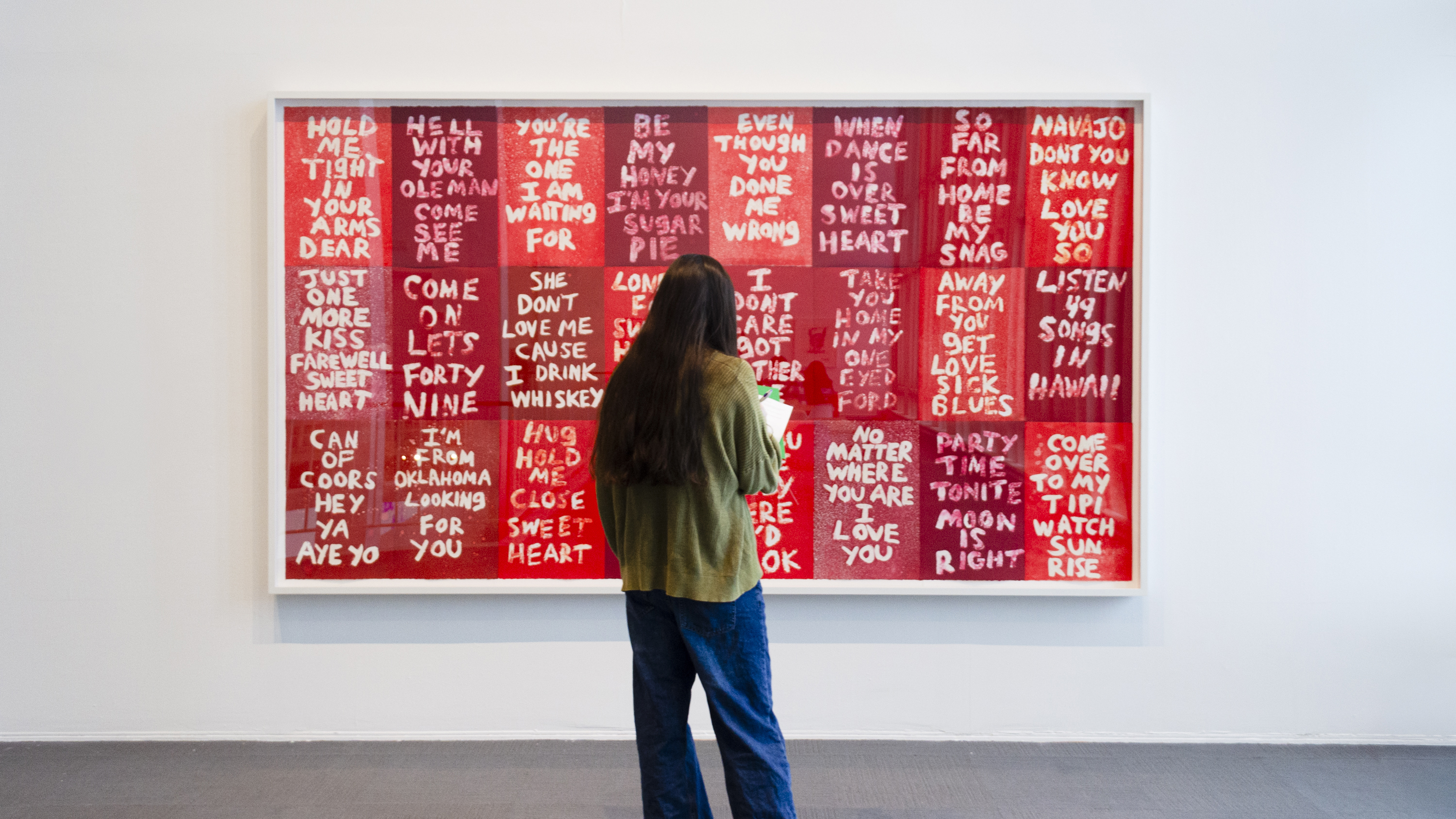 A university student stands and looks intently at a large red colorful artwork with white text by Hock E Aye Vi Edgar Heap of Birds that was included in the exhibition History Lessons. 