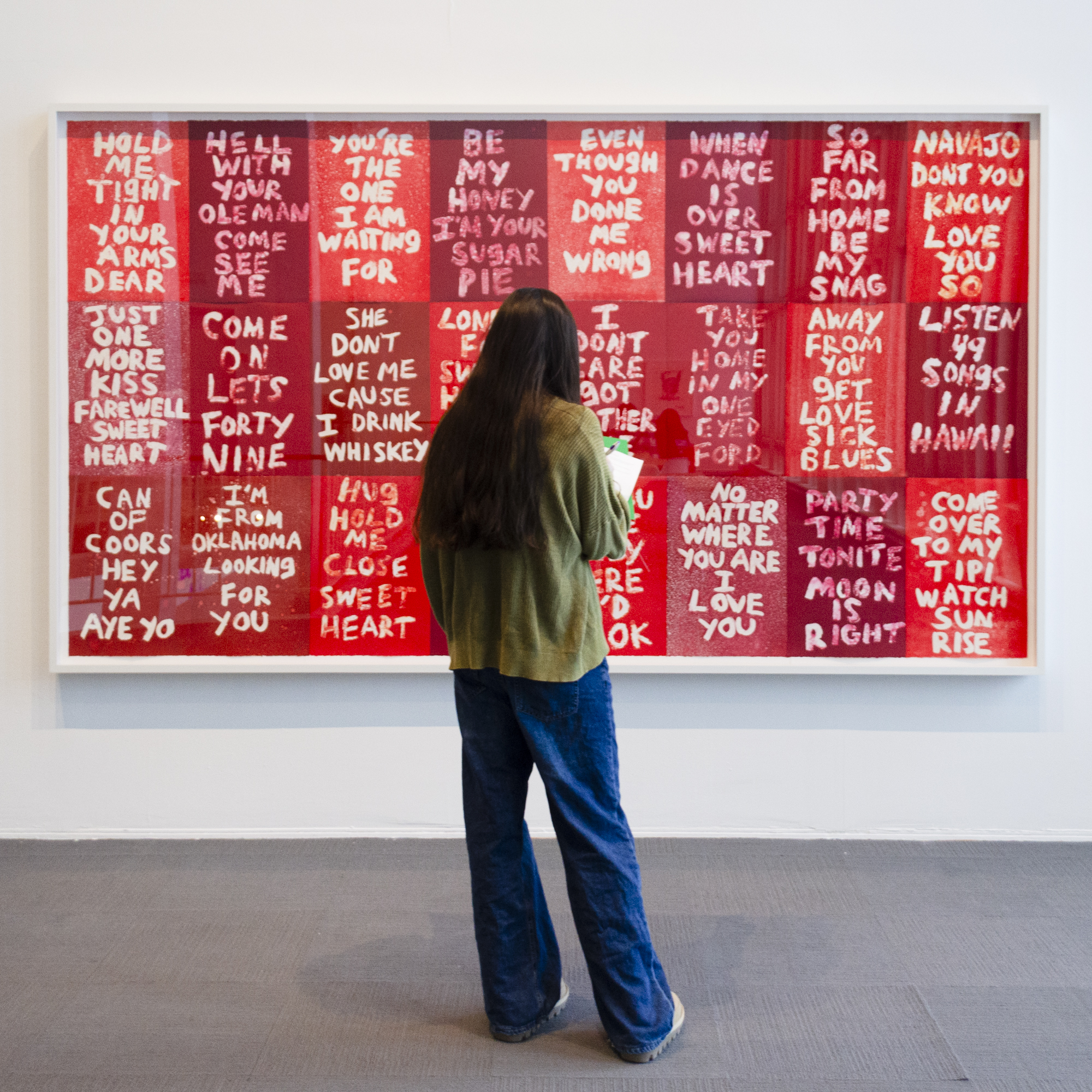 A university student stands and looks intently at a large red colorful artwork with white text by Hock E Aye Vi Edgar Heap of Birds that was included in the exhibition History Lessons. 
