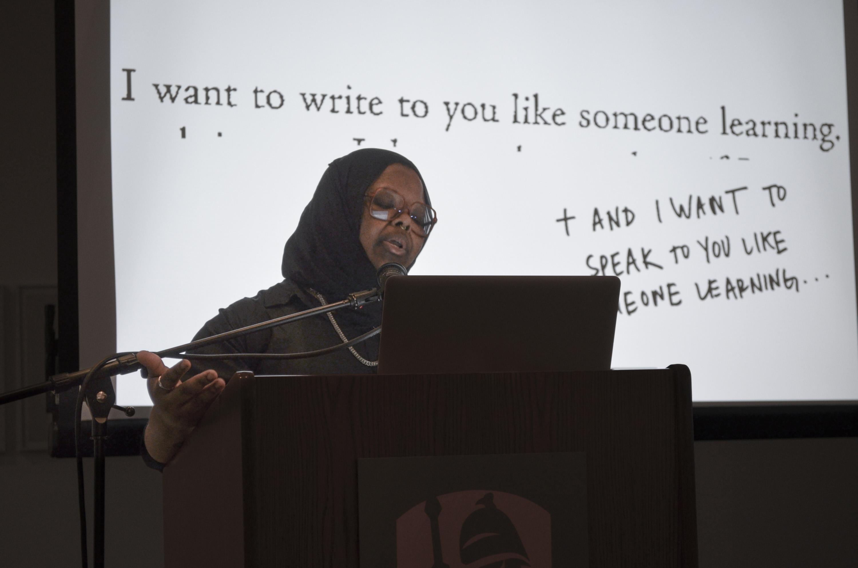Kameelah Janan Rasheed stands behind a podium, with the backdrop of a brightly lit projector screen for a public program on the first floor of the University Art Museum. Text on the projected image reads I want to write to you like someone learning and I want to speak to you like someone learning.