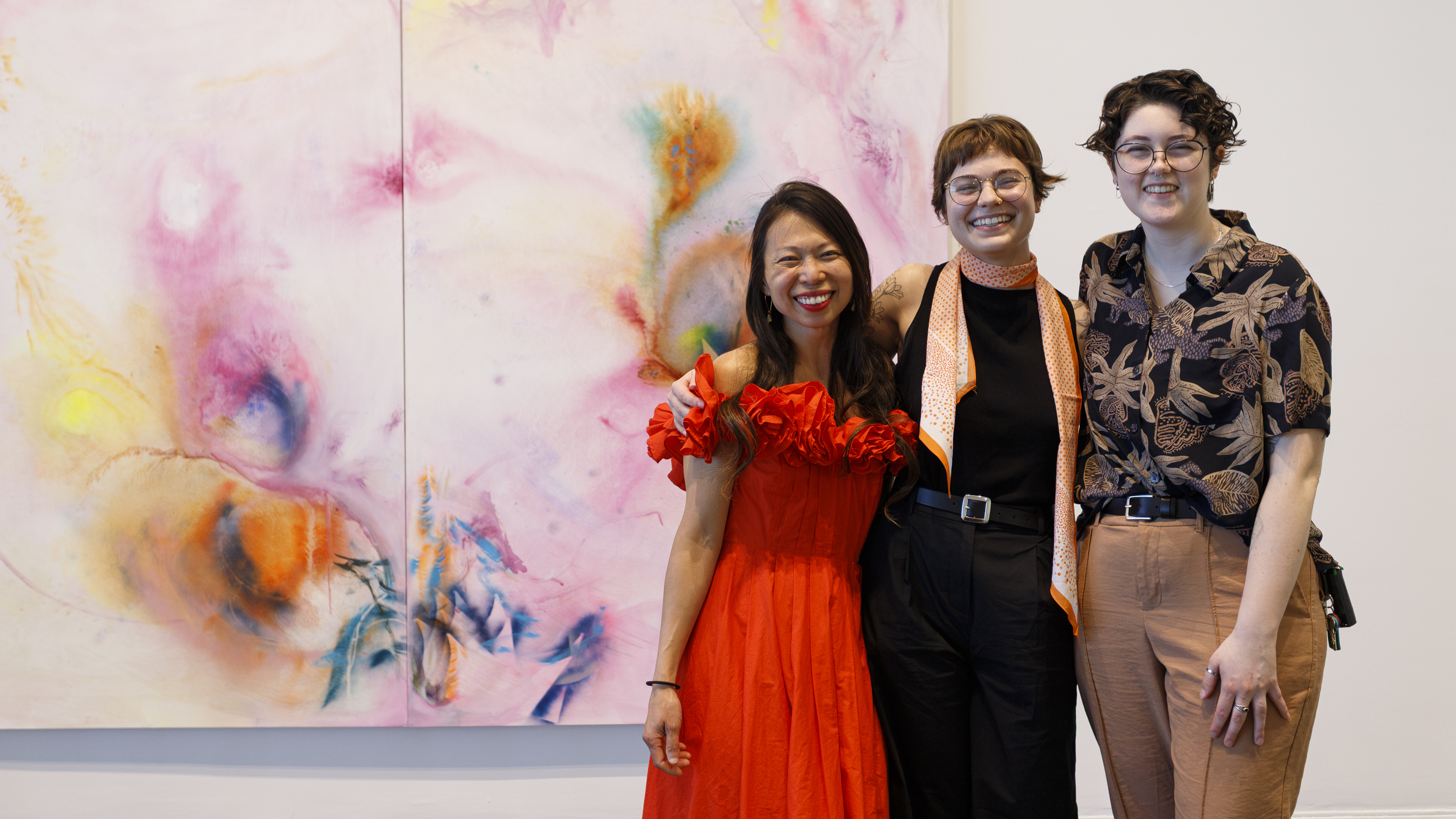 MFA graduate students Kim Tateo, Ally DeRusso, and Avery Hartranft stand smiling in front of Tateo’s large colorful painting. 