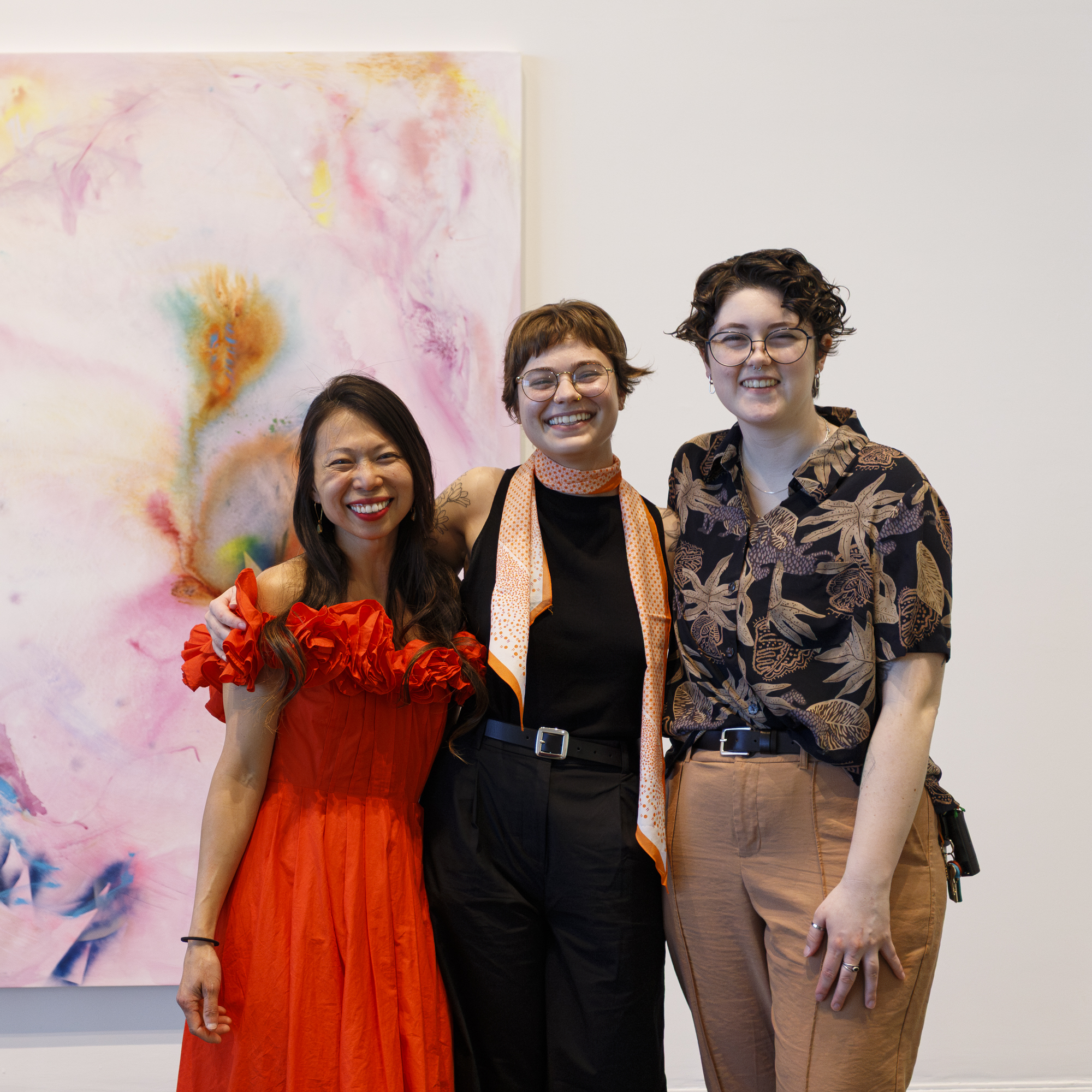 MFA graduate students Kim Tateo, Ally DeRusso, and Avery Hartranft stand smiling in front of Tateo’s large colorful painting. 