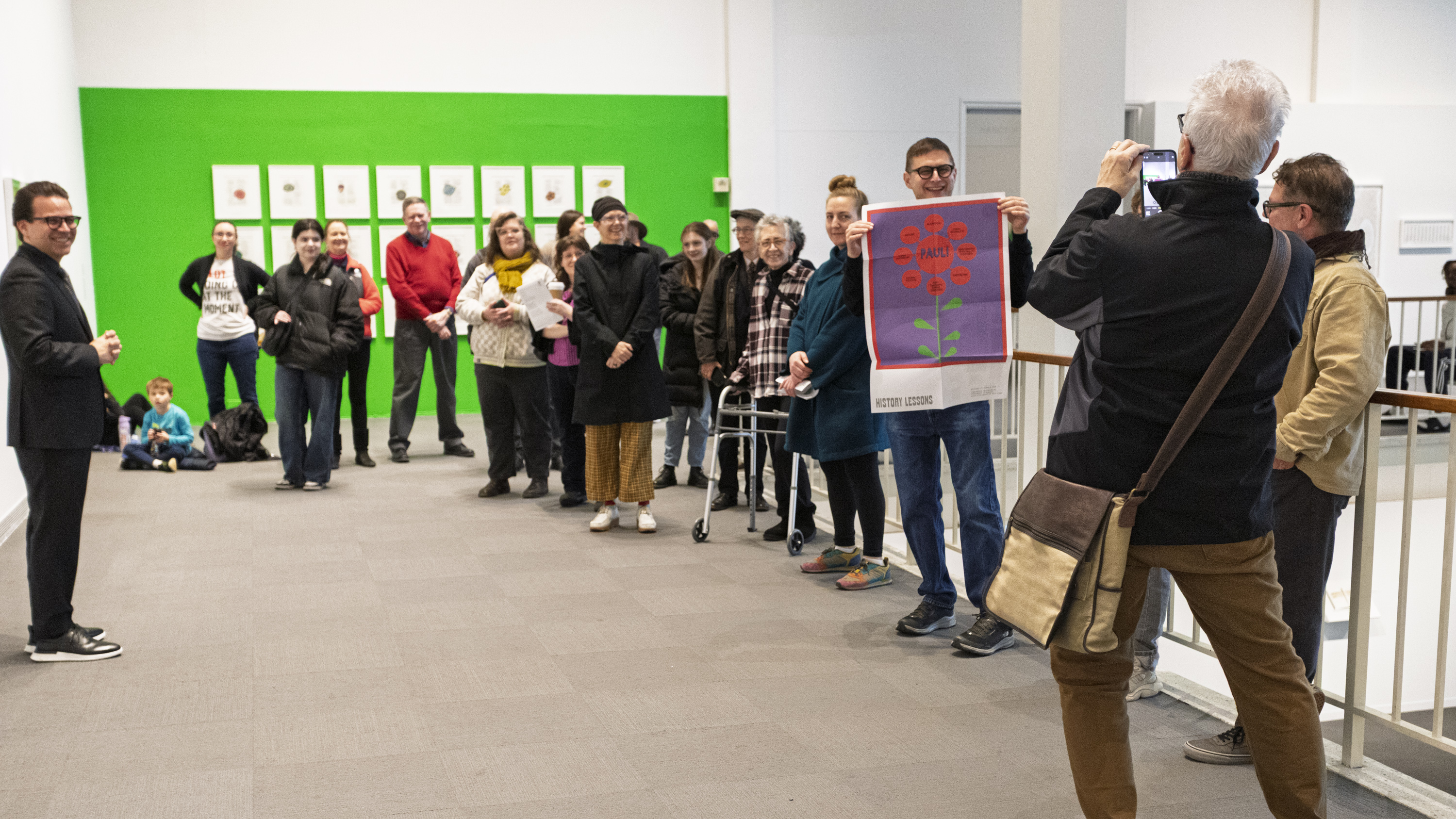 A large group of visitors on the second floor of the University Art Museum are laughing and smiling as exhibiting artist Joe Mama Nitzberg holds up the red colorful flower-like reproduction of this artwork in the exhibition brochure. 