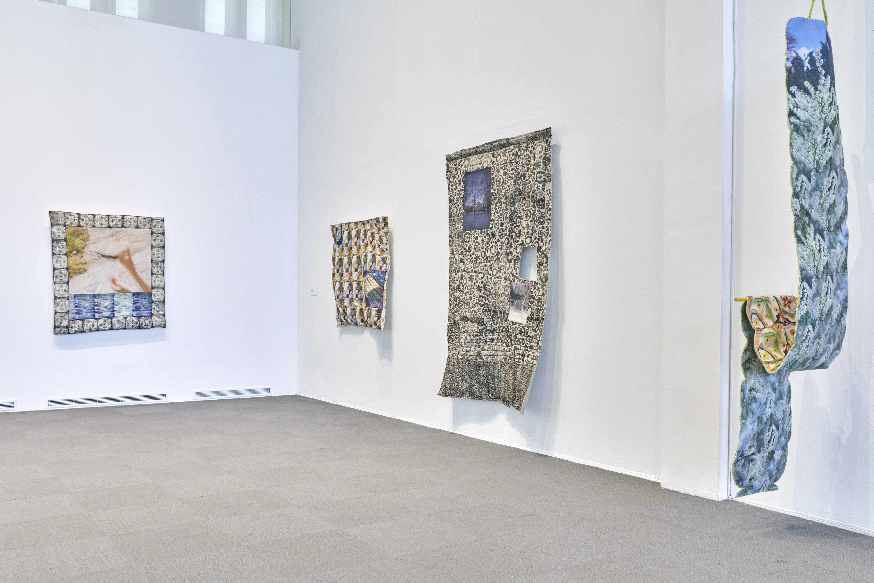 Four large textile artworks are hung on the white walls of the University Art Museum. The textiles are made up of earth tones and blue-green hues. 