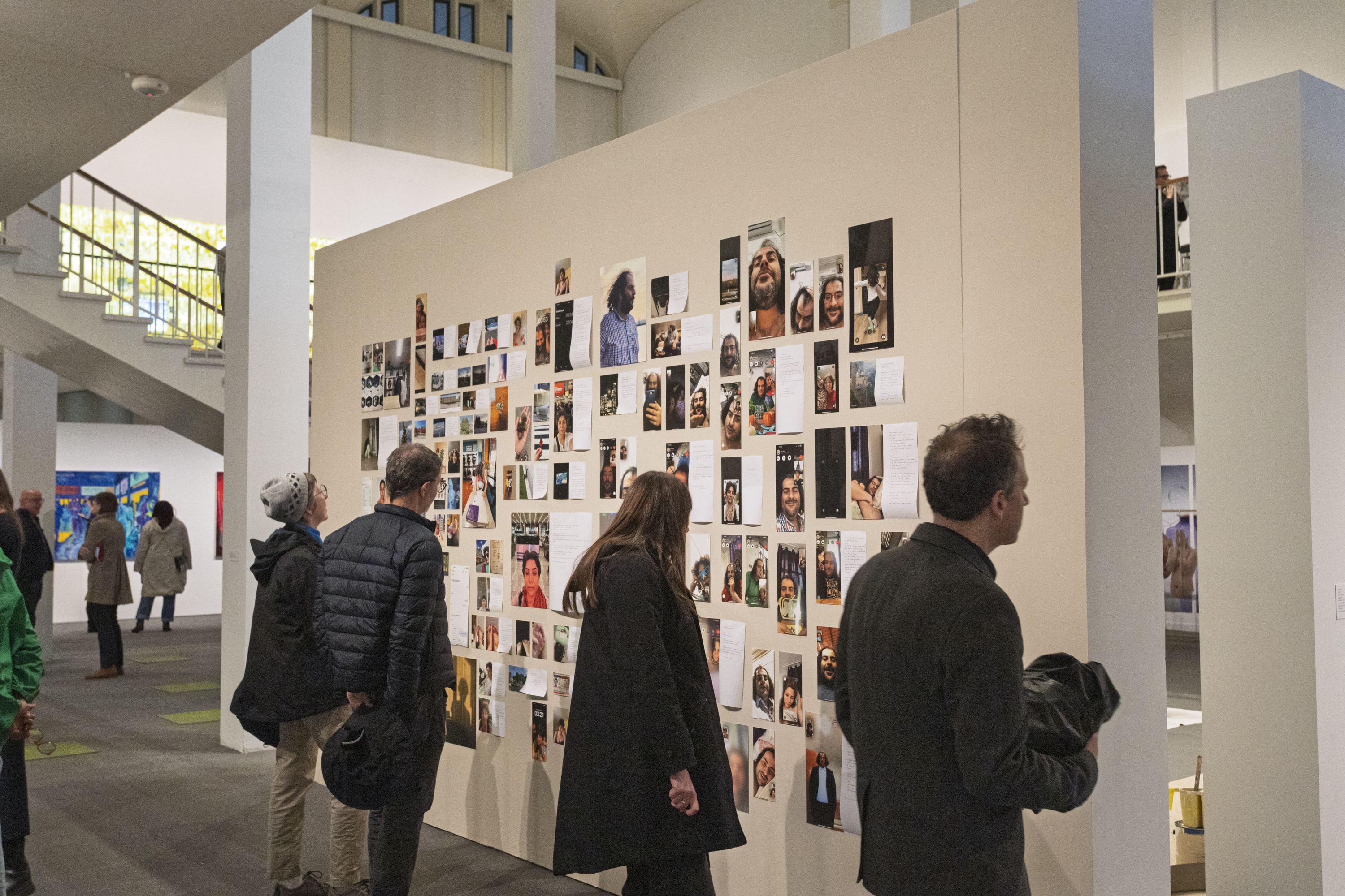 Visitors are walking around the first floor of the University Art Museum and are looking closely at a wall full of photographs and text by exhibiting MFA artist Hoora Mirabzadeh.