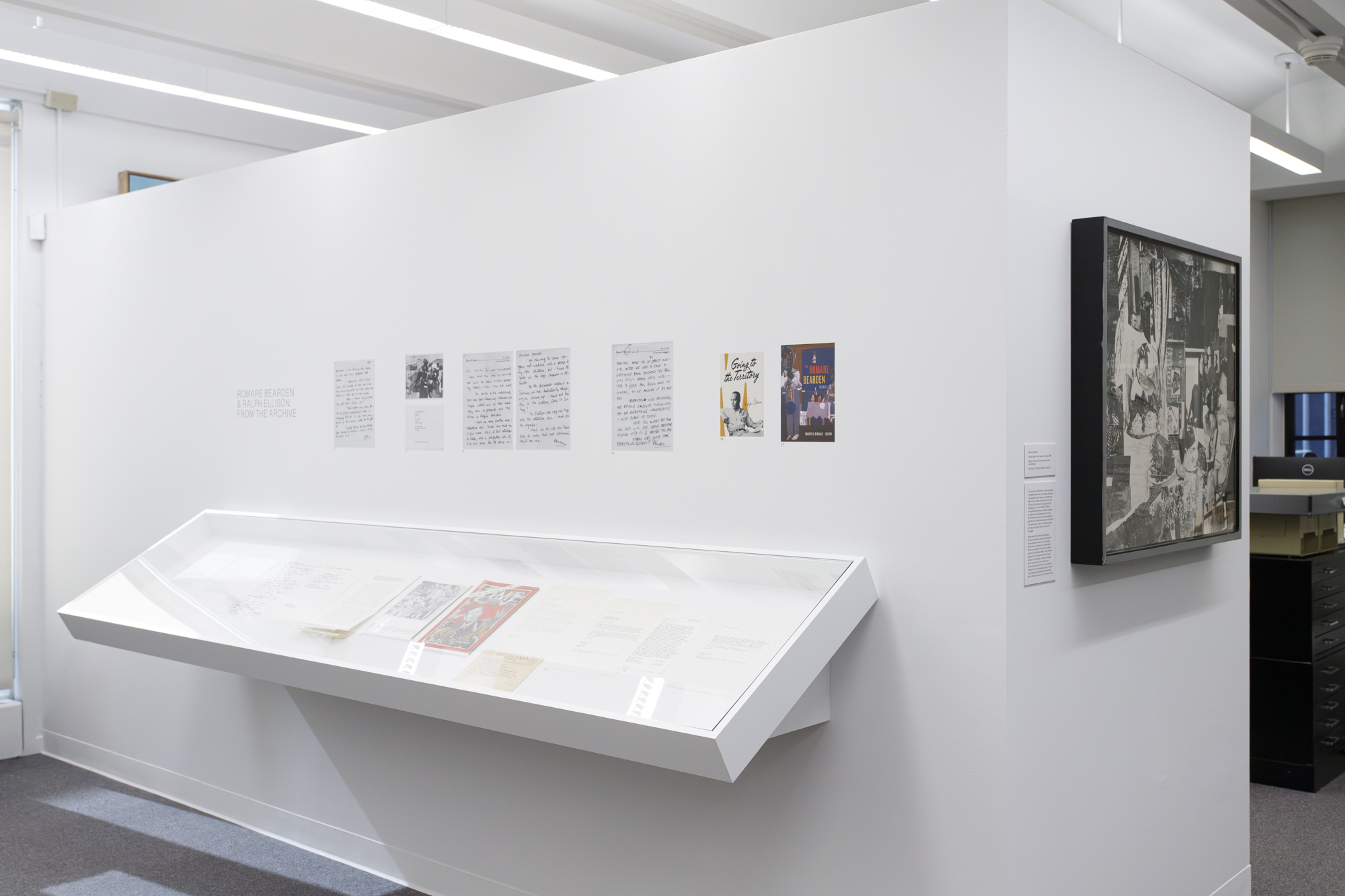 Black and white handwritten letters and typed documents sit within a display case mounted to a white wall. Vinyl reproductions of correspondence are installed on the wall above the case, and a black and white artwork by Romare Bearden is installed on the adjacent wall.