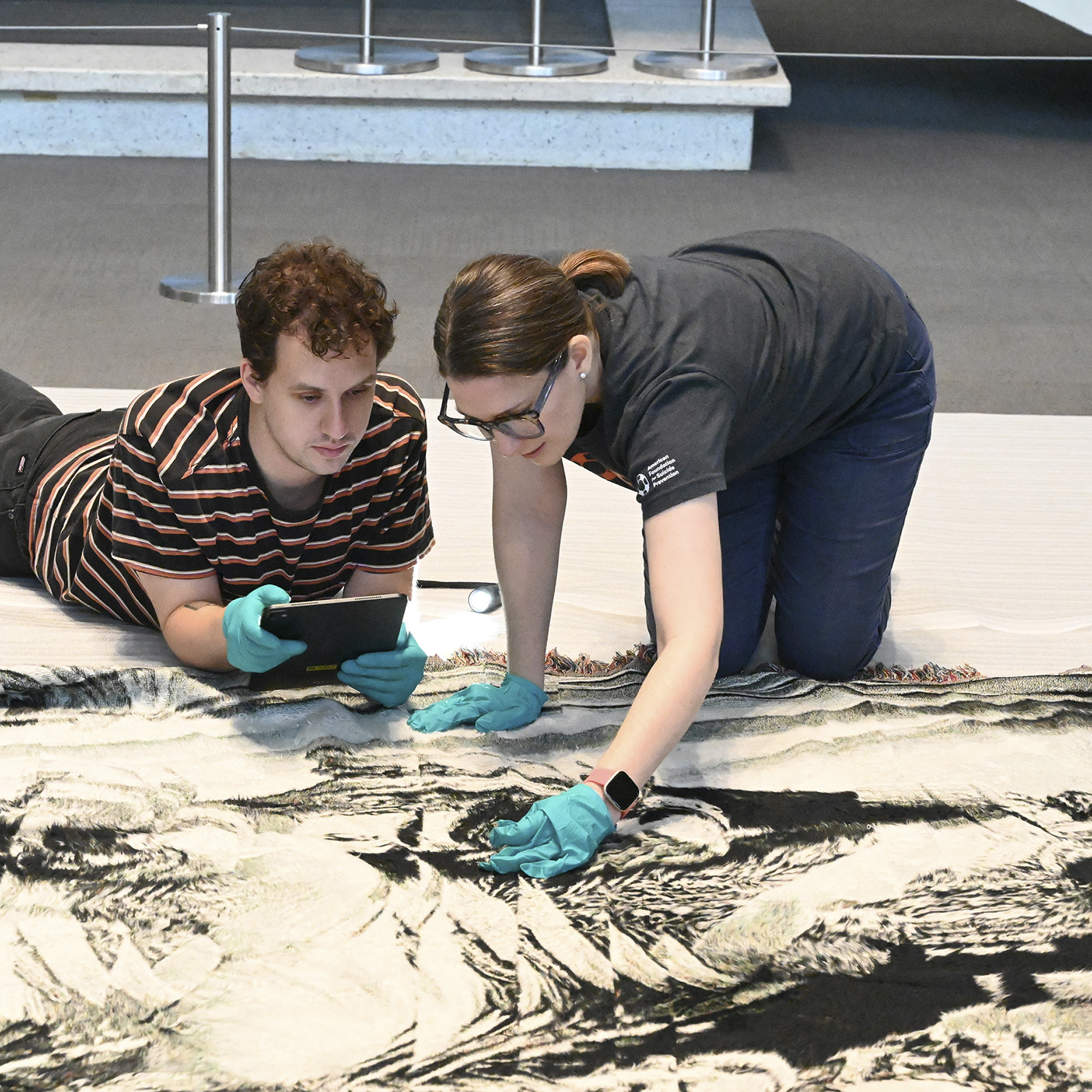 Christian Henry Wechgelaer MFA ’25, Museum Assistant, and Darcie Abbatiello, Registrar/Collections Manager, wear aqua nitrile gloves and closely examine a black and white tapestry by exhibiting artist Noel W Anderson.